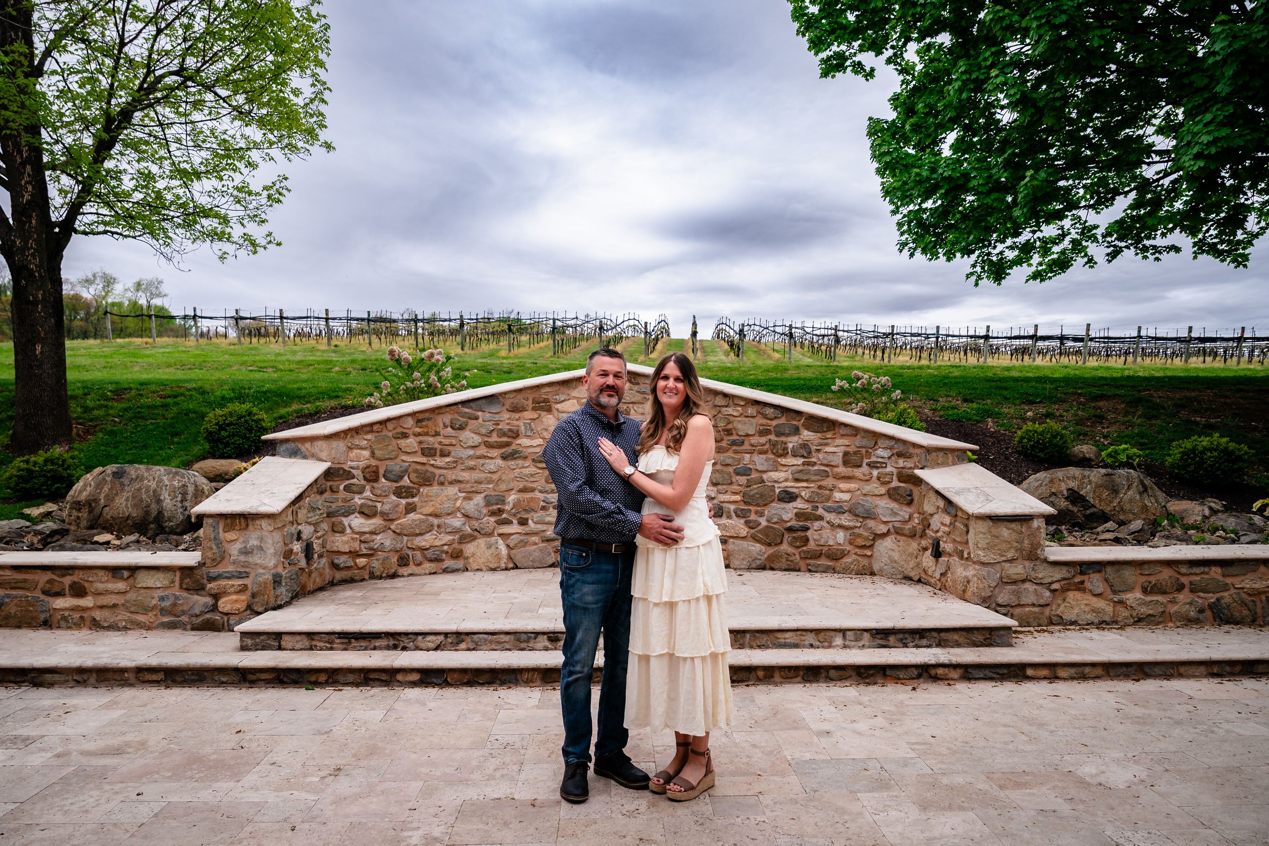 Couple embracing in front of a stone wall at a vineyard wedding venue under cloudy skies.