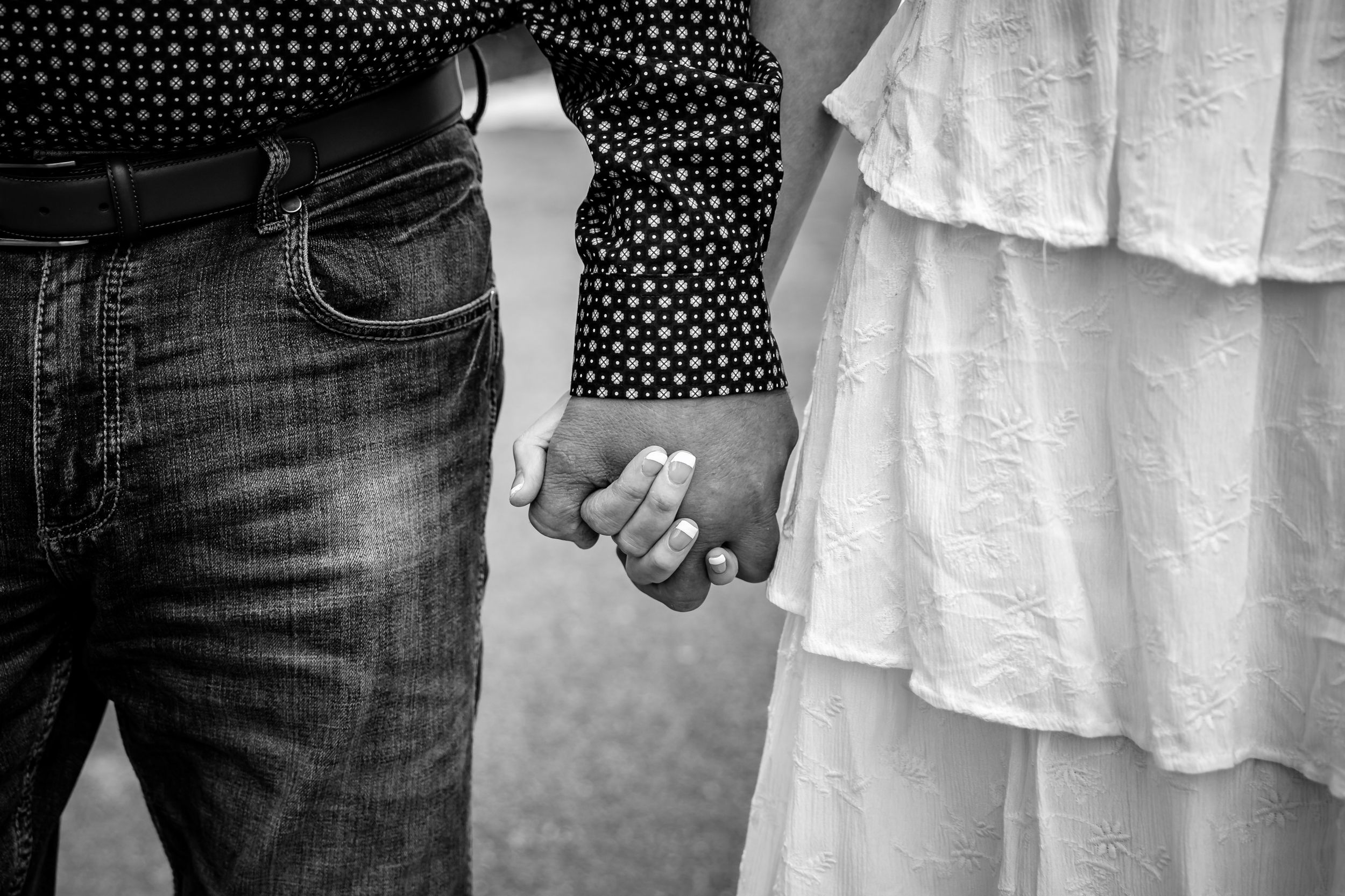 Black and white photo of a couple holding hands, woman wearing white dress and engagement ring, man in jeans.