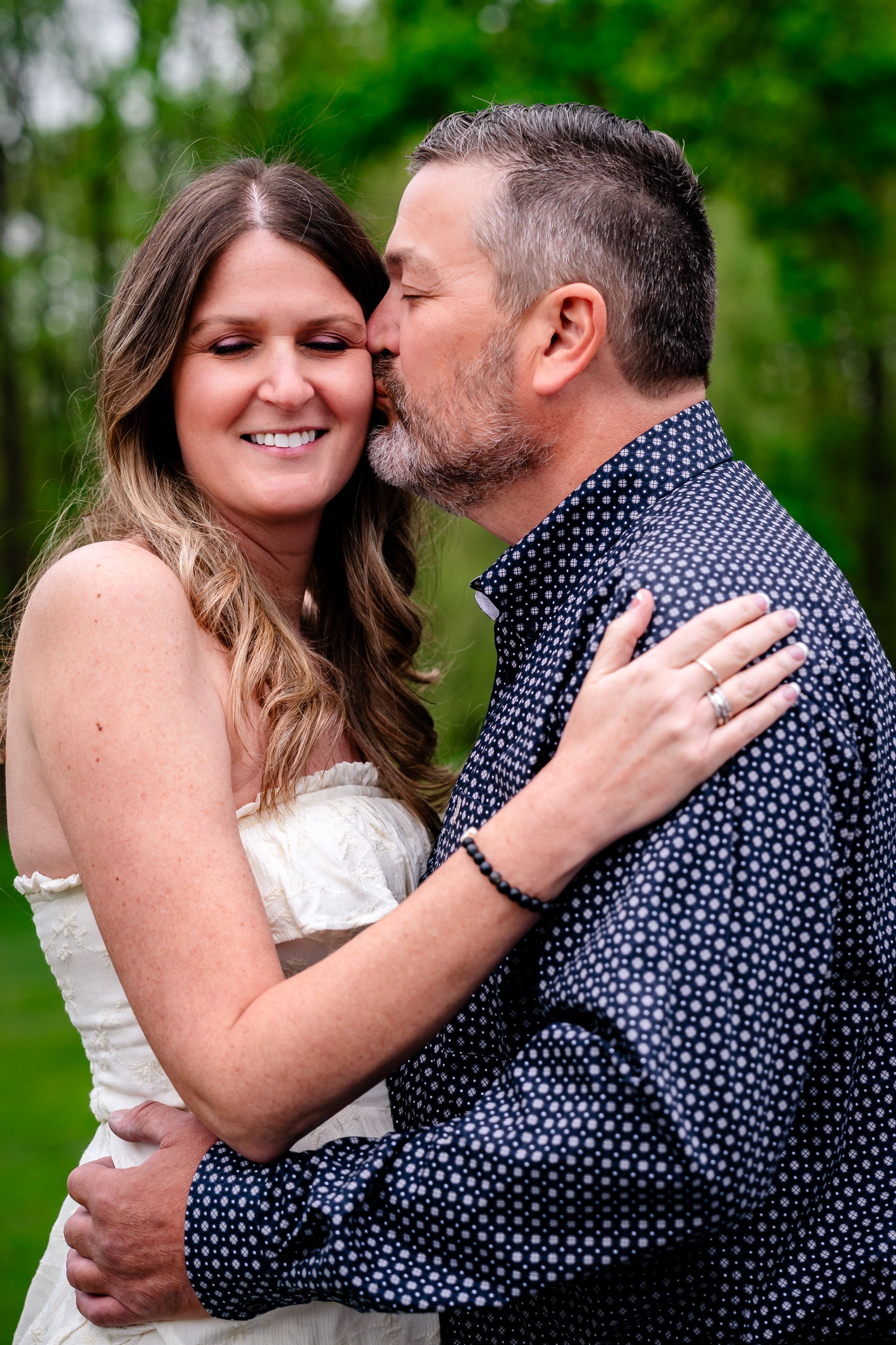Groom kissing bride on cheek as she smiles during romantic outdoor engagement photo session.