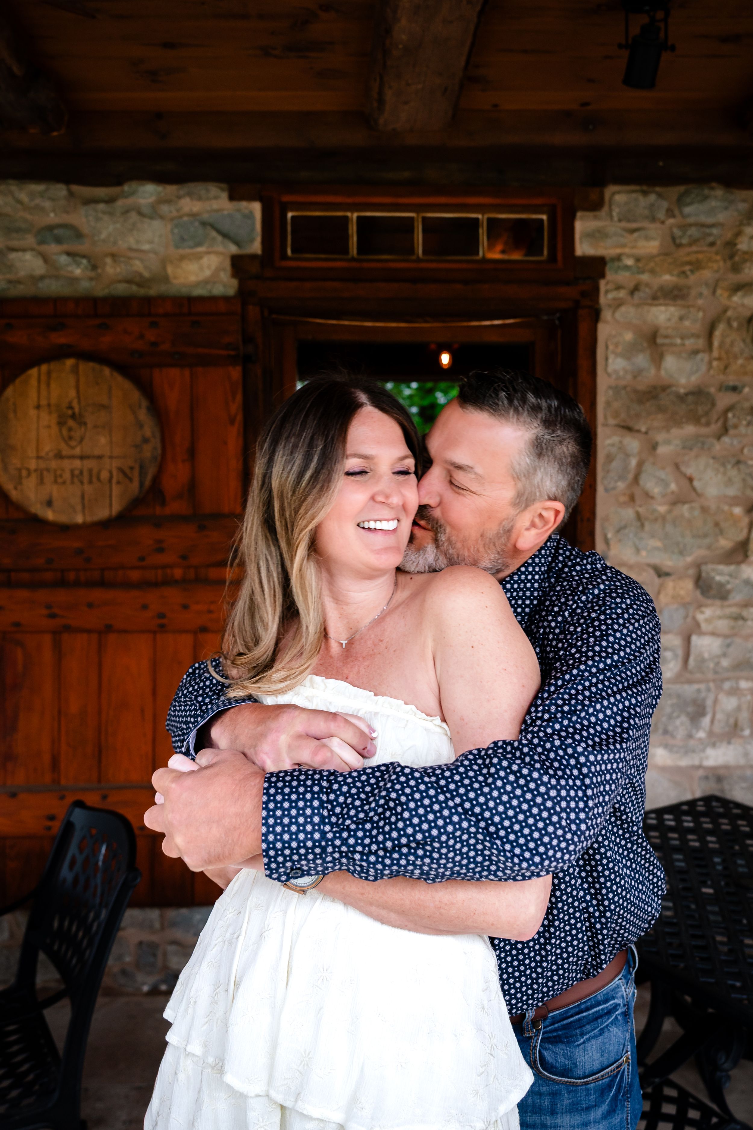 A couple embraces and laughs outside a rustic stone building, the man kissing the woman's cheek as she smiles joyfully.