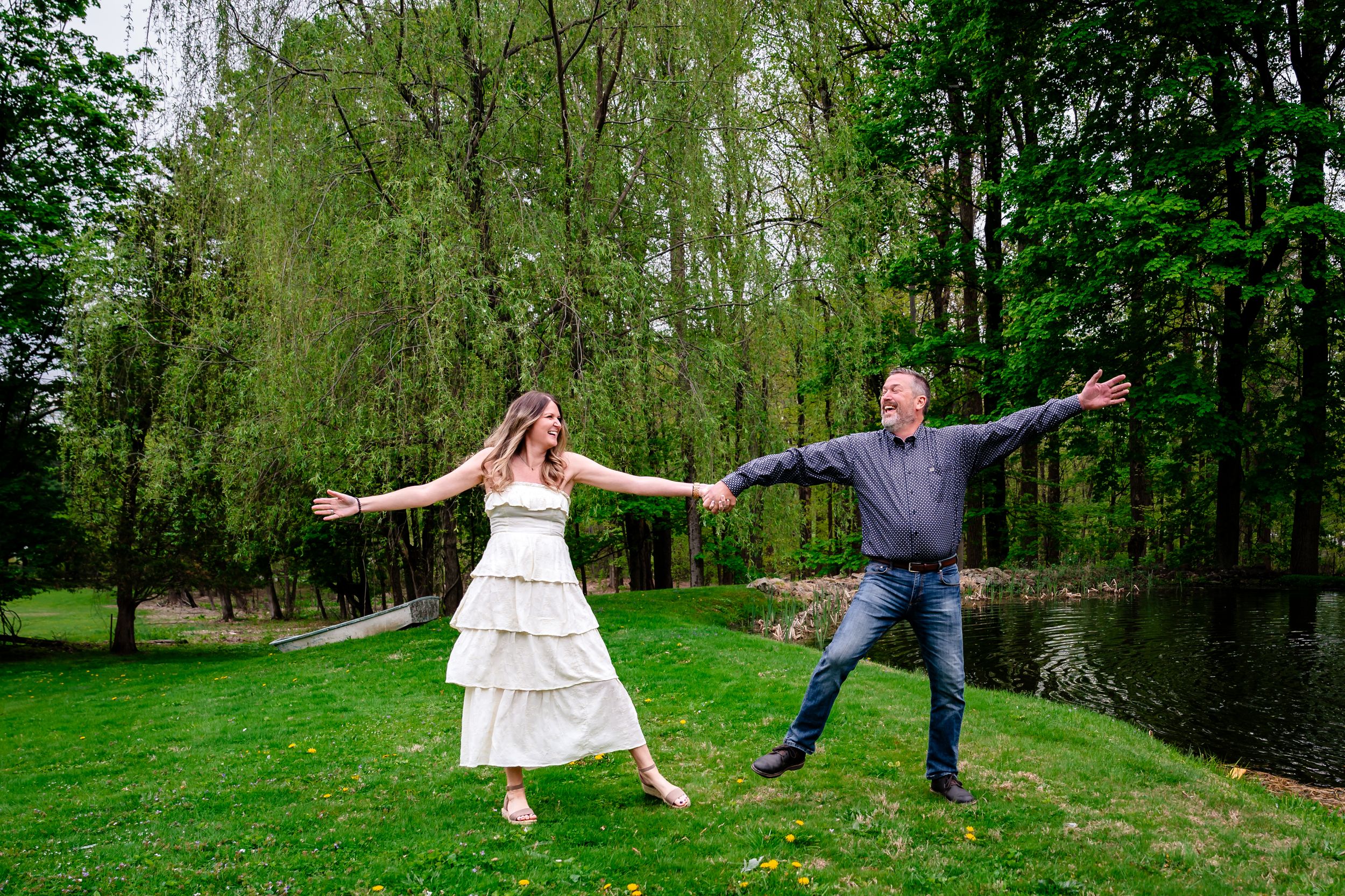 Joyful couple holding hands and dancing outdoors on green grass with willow trees and a pond in the background.