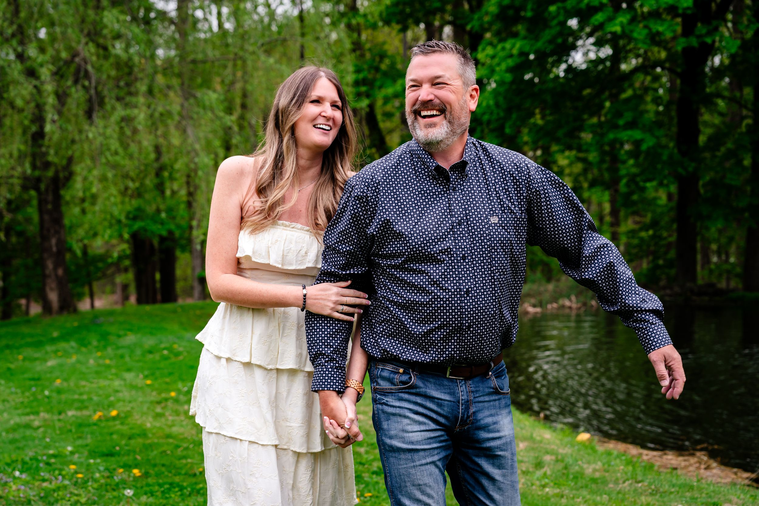 Smiling couple holding hands outdoors near a pond, woman in white dress and man in dark shirt, surrounded by green trees.