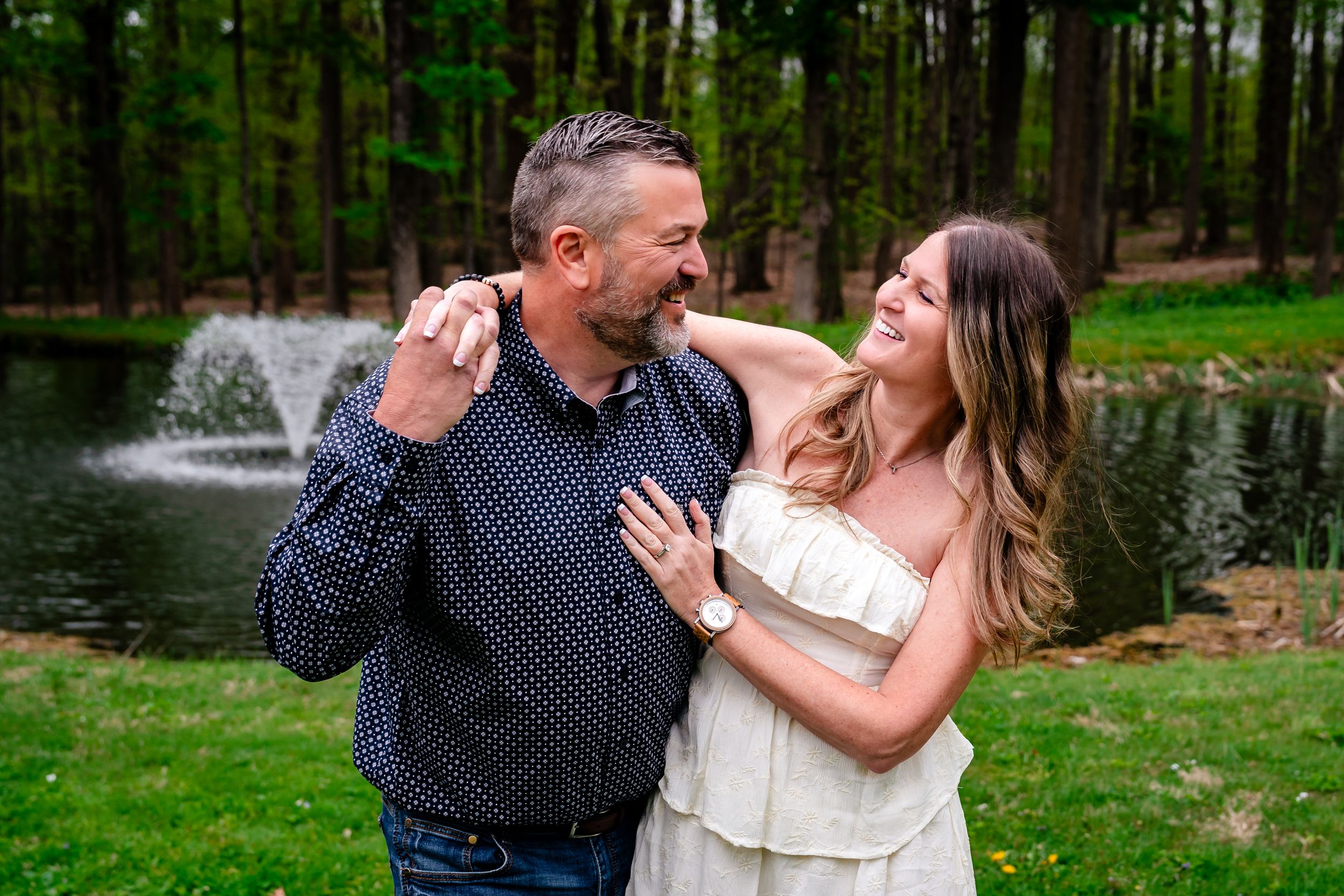 Happy couple embracing and smiling by a pond with fountain surrounded by trees in an outdoor setting.