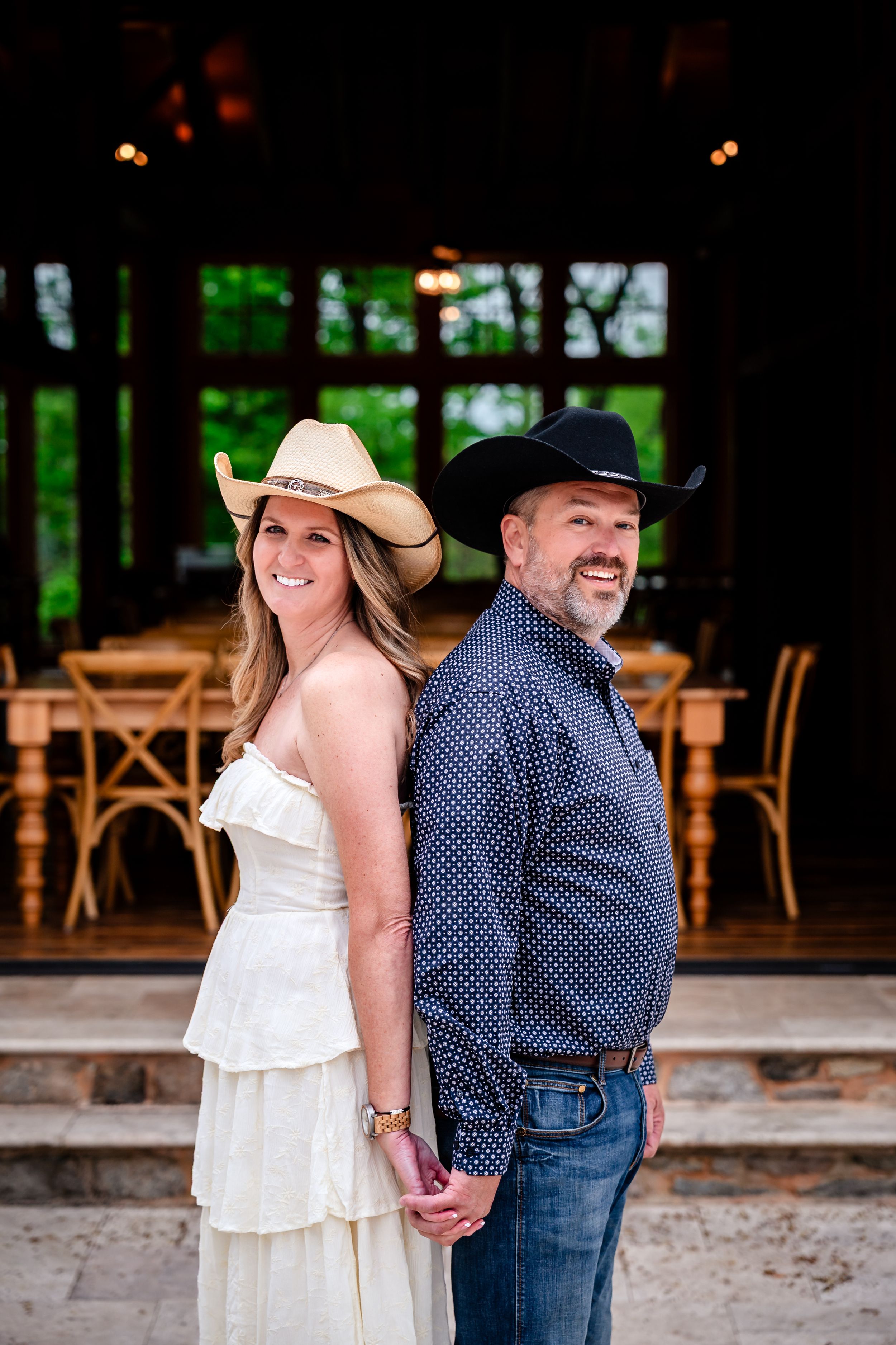 Couple in cowboy hats holding hands back-to-back, woman in white dress, man in navy shirt, rustic barn venue.