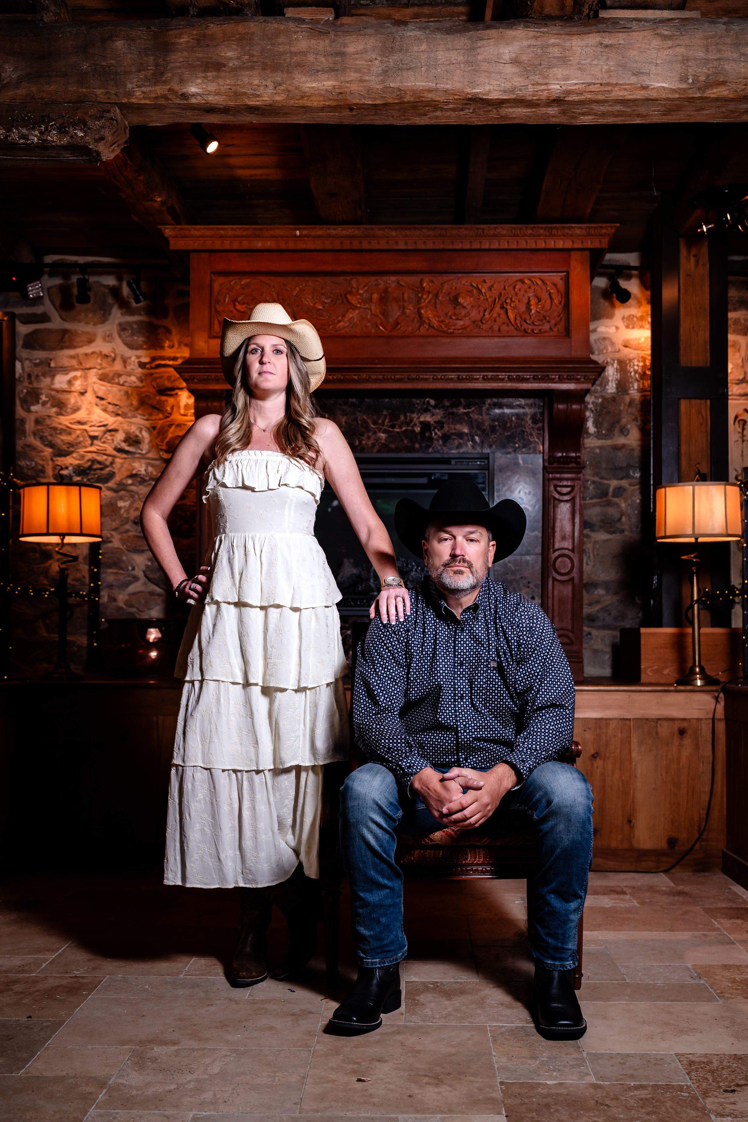 Couple posing in rustic lodge, woman standing in white tiered dress with cowboy hat, man seated in jeans.