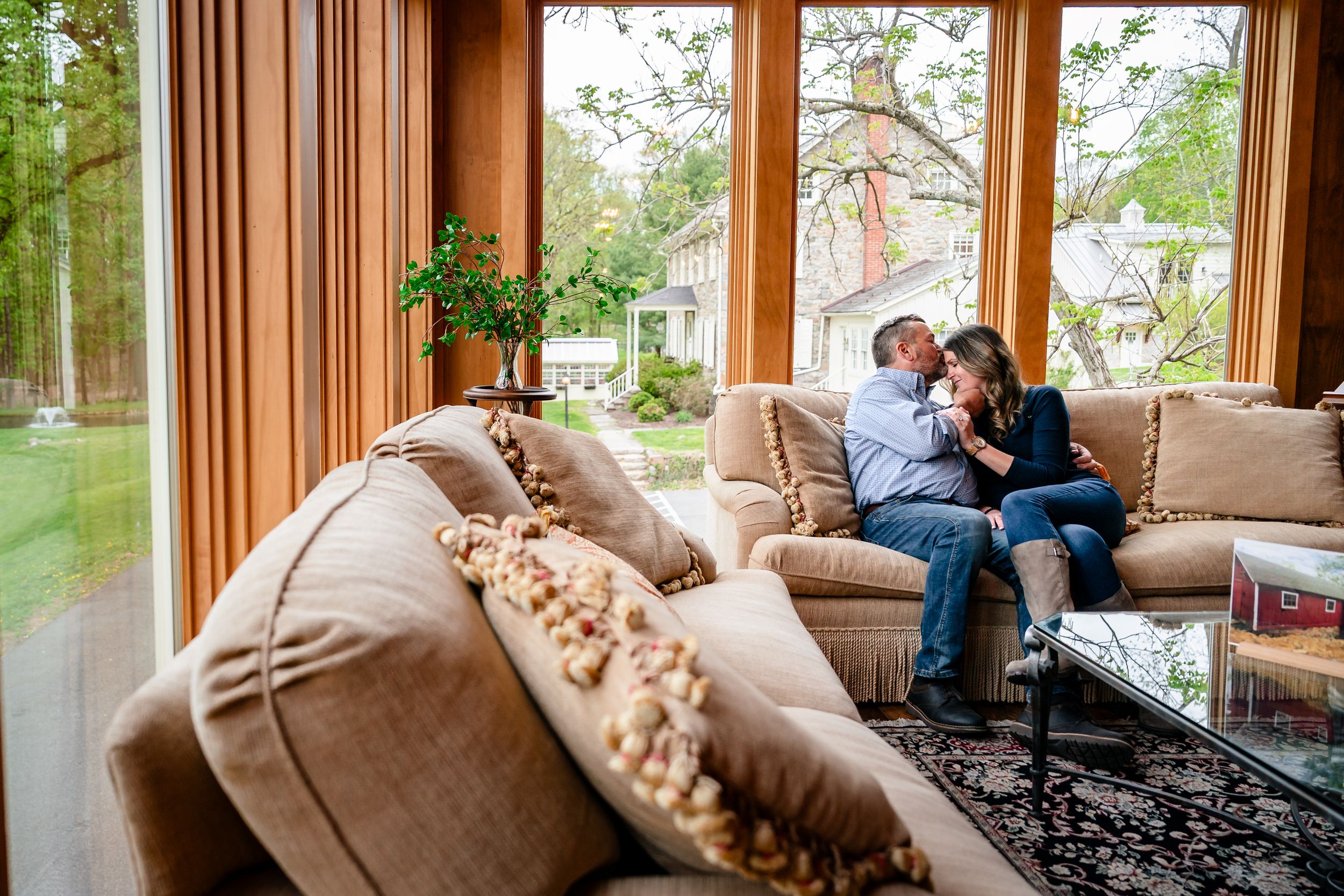 Couple sharing a tender moment on a beige sofa in a sunlit living room with large windows overlooking a garden.
