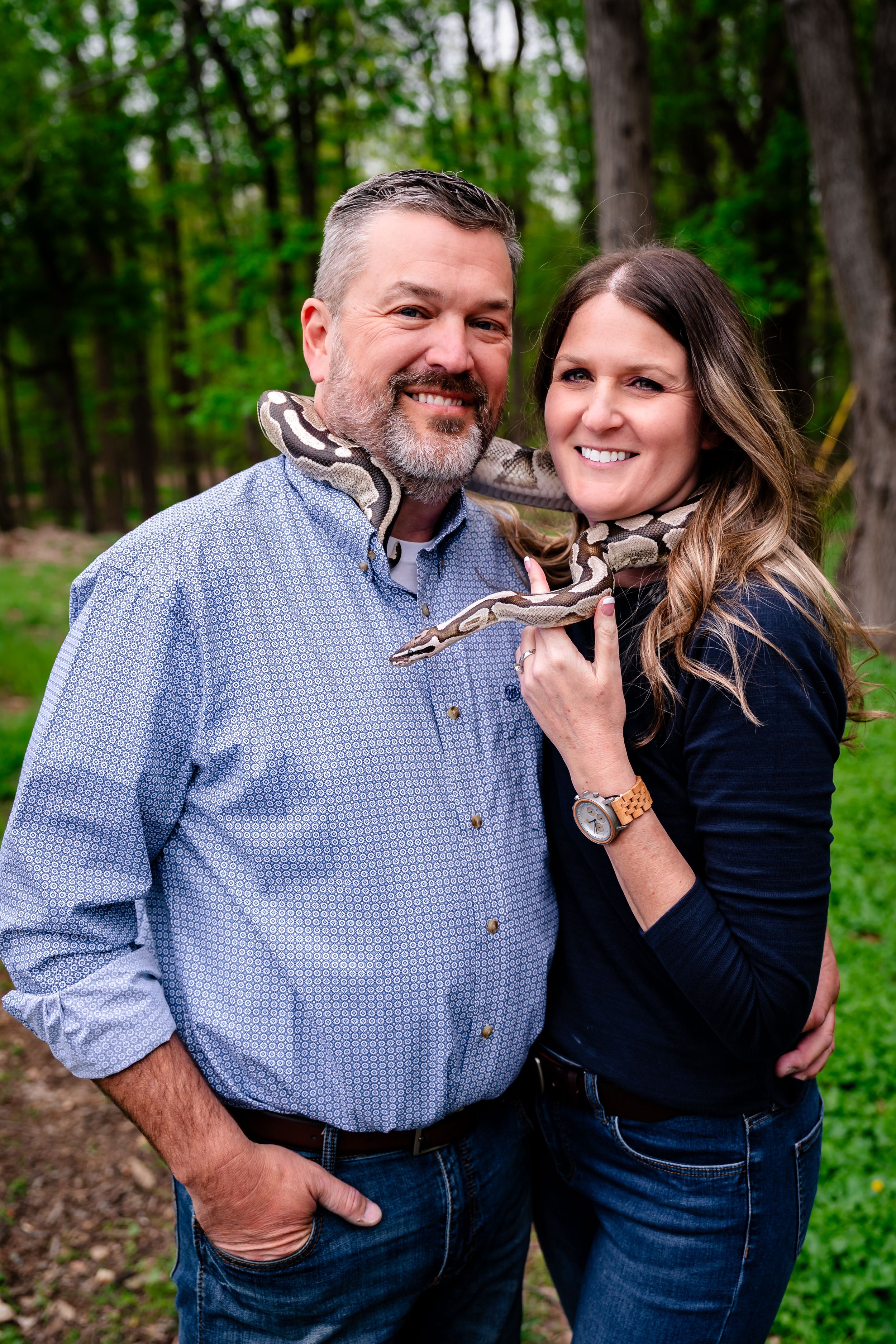 Smiling couple posing outdoors with a large boa constrictor snake draped around their shoulders in a wooded setting.