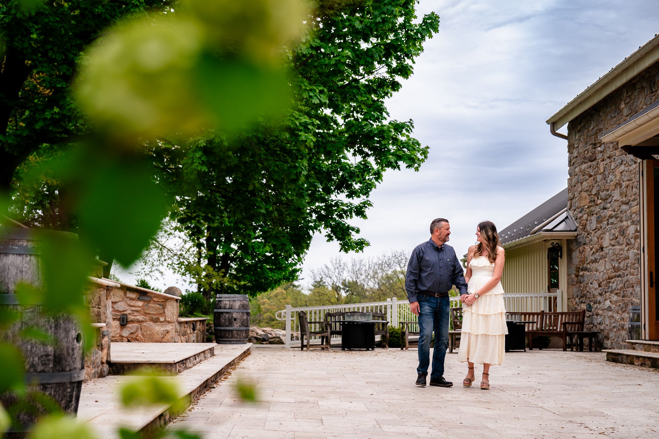 Couple holding hands on a stone patio beside a rustic building surrounded by lush green trees.