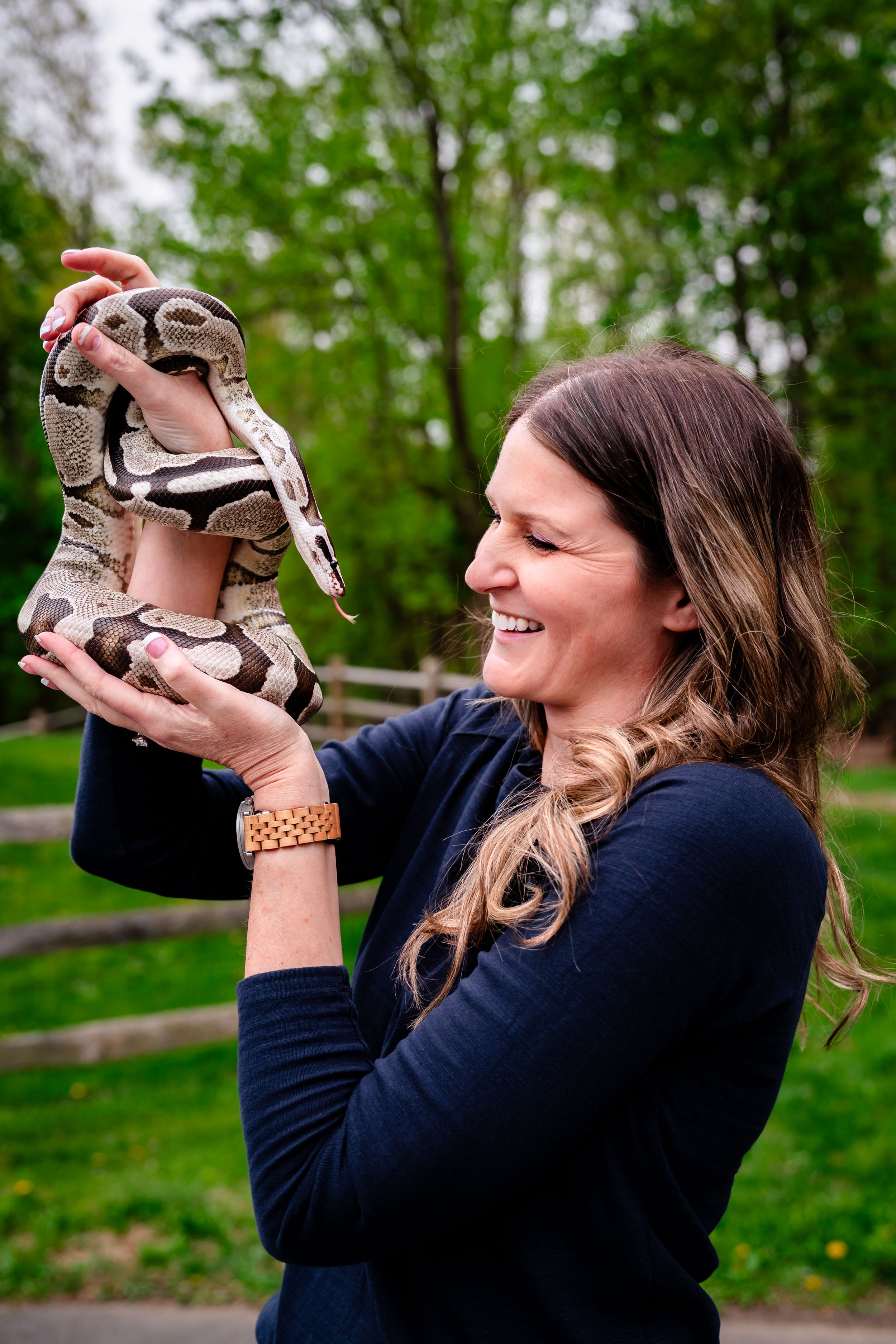Smiling woman in navy shirt holds a ball python snake outdoors with green trees in background.