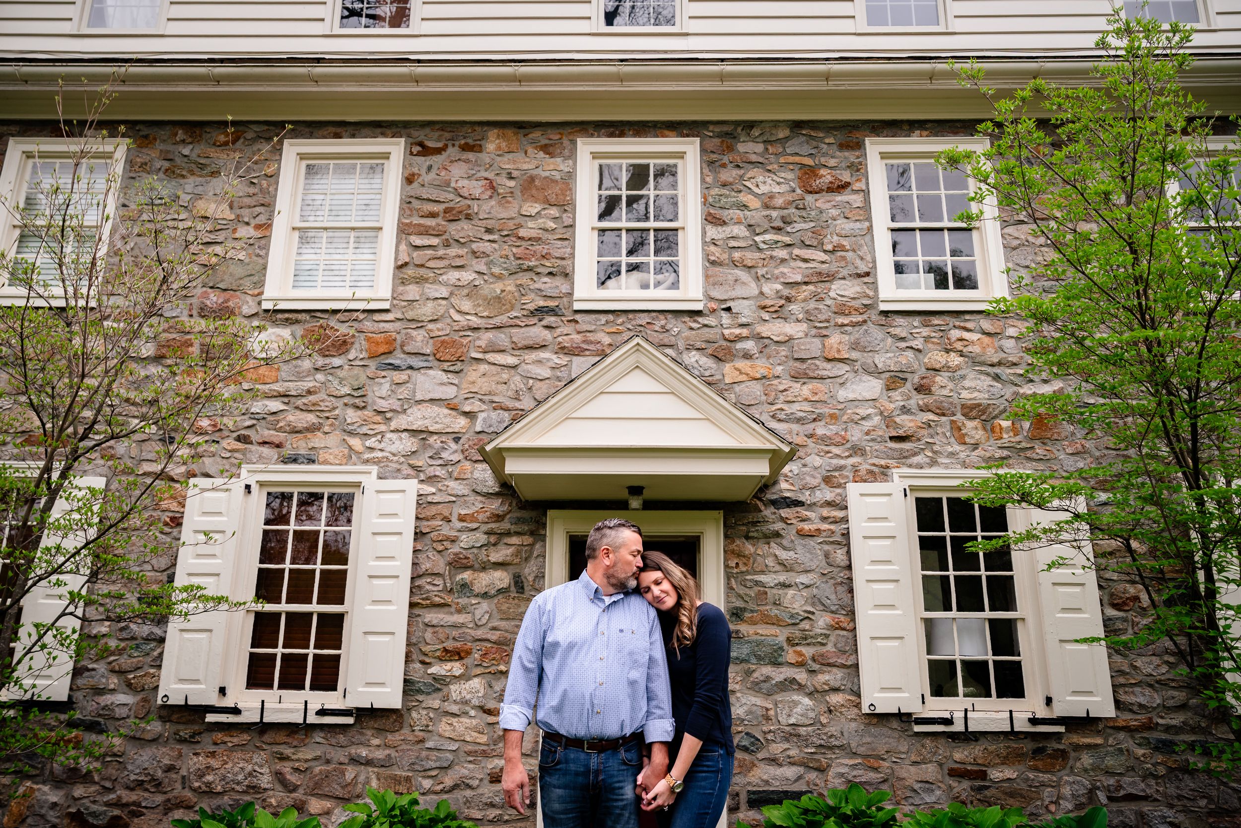 Couple embracing in front of a historic stone house with white shutters and a classic pediment doorway.