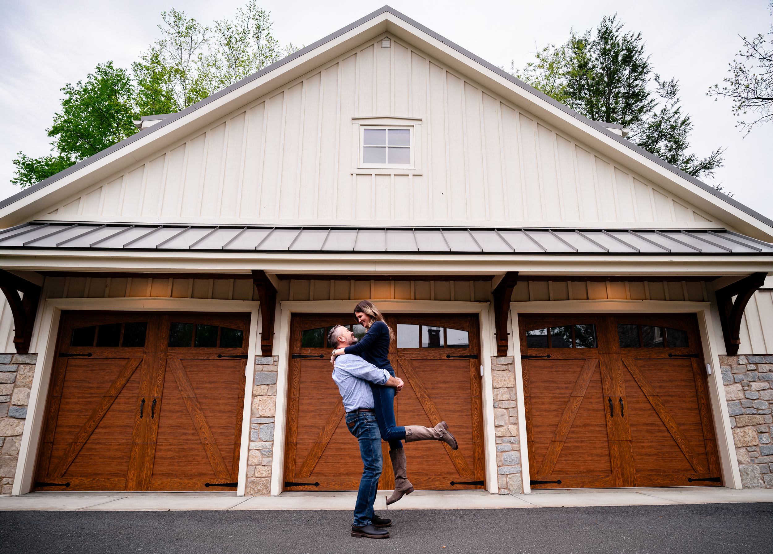 Couple sharing a romantic lift embrace in front of a charming barn-style garage with wooden doors.