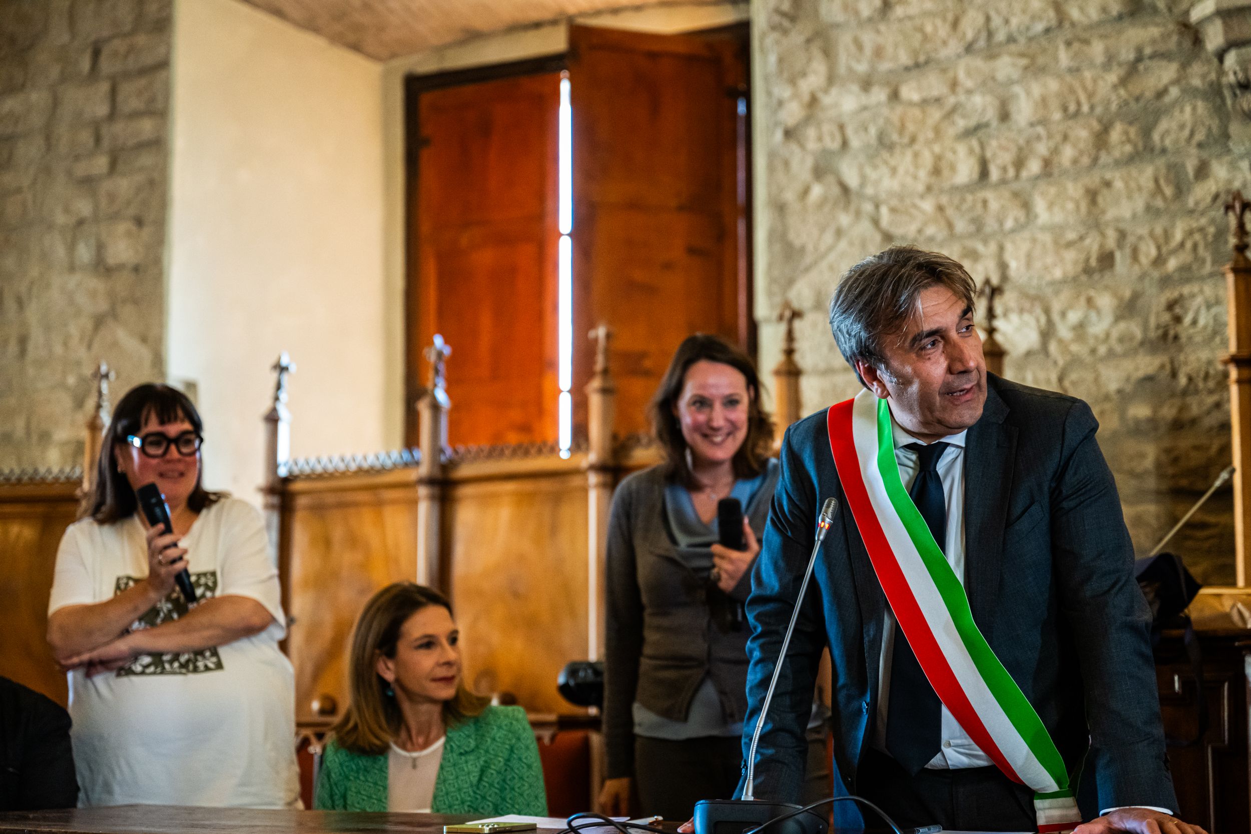 Italian mayor in sash speaks at indoor ceremony with attendees in historic stone building.