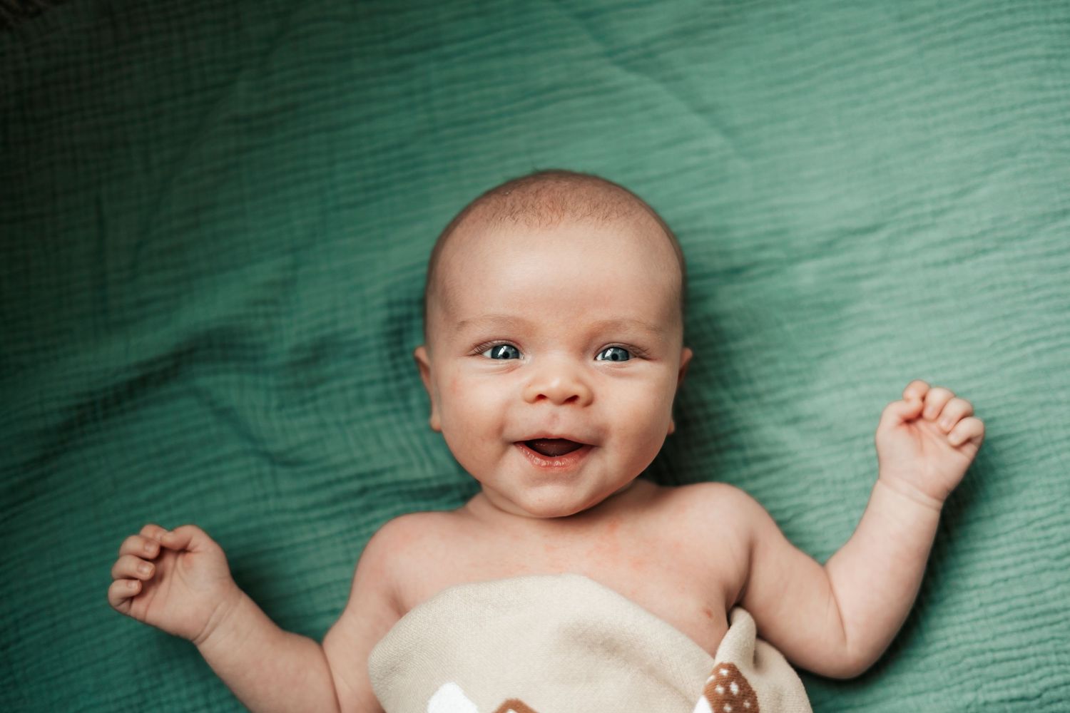 Smiling newborn baby lying on a teal blanket, wrapped in a cream swaddle  with bright eyes  | Newborn Photographer in Broomfield Colorado