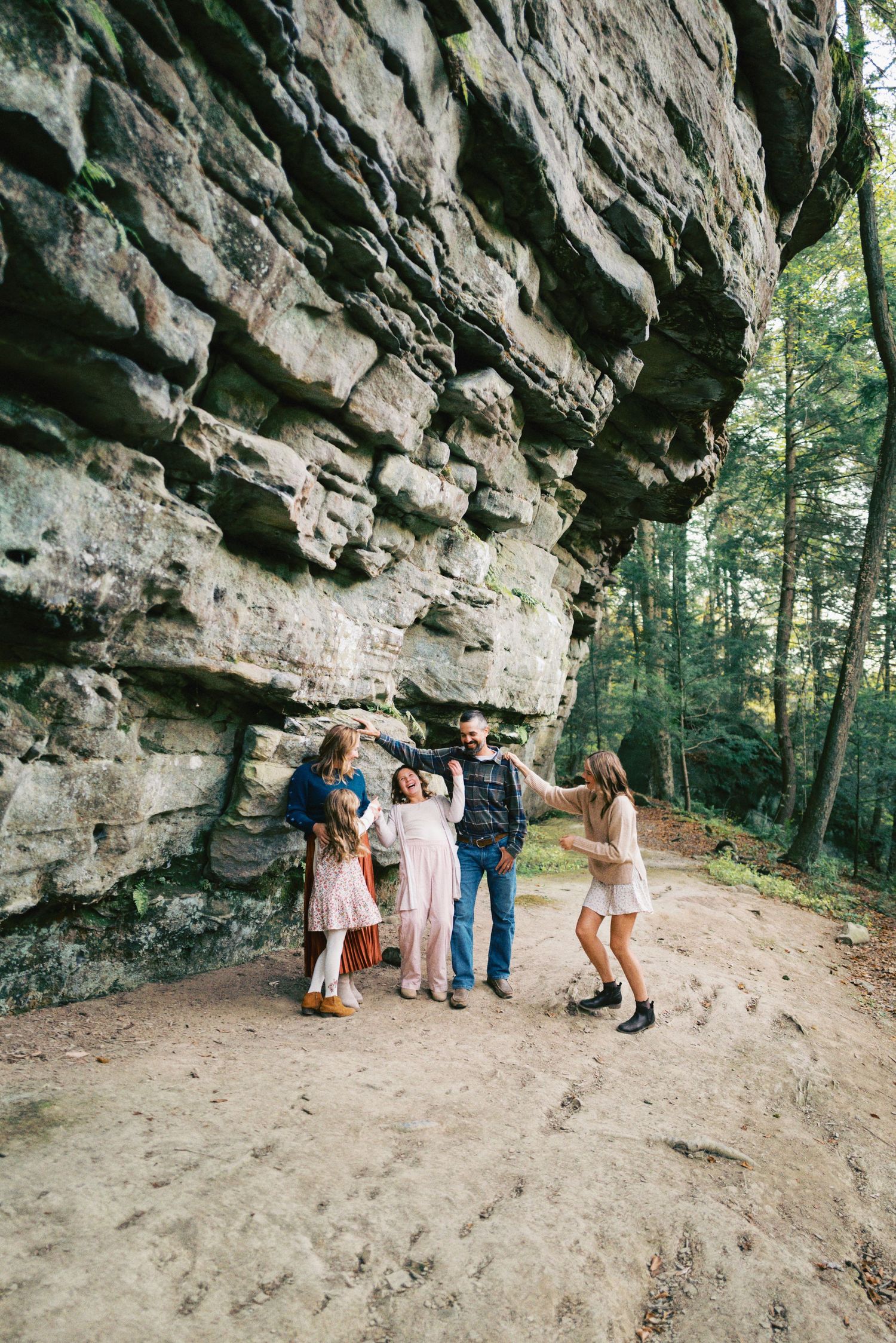 Family of five posing and dancing near a large rock formation in a forest setting.