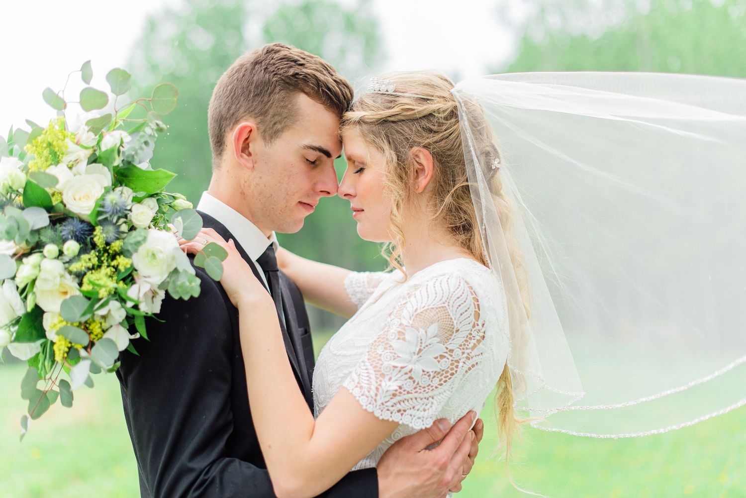 A bride and groom share an intimate moment, touching foreheads, as her veil flows in the breeze during their wedding day.