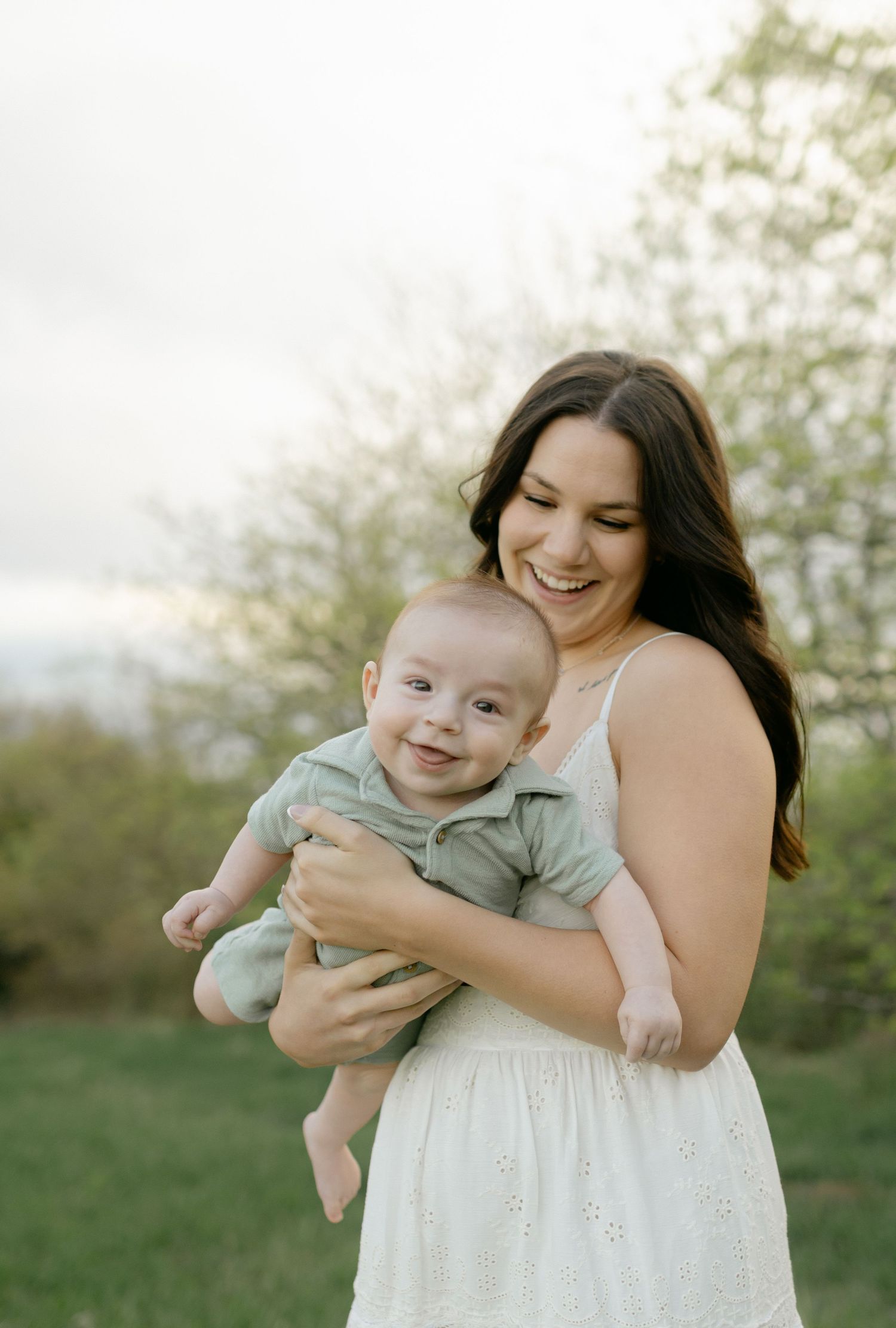 Joyful brunette mother in white dress laughs while holding her smiling baby boy outdoors at dusk.
