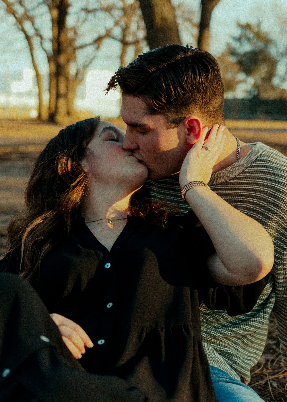 A couple sharing a romantic kiss outdoors in a sunlit park setting.