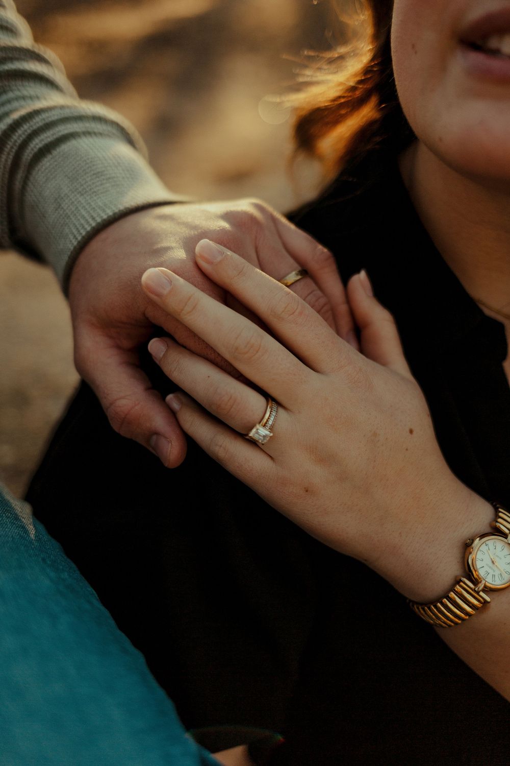 Close-up of an engaged couple holding hands, showcasing a diamond engagement ring and gold watch.