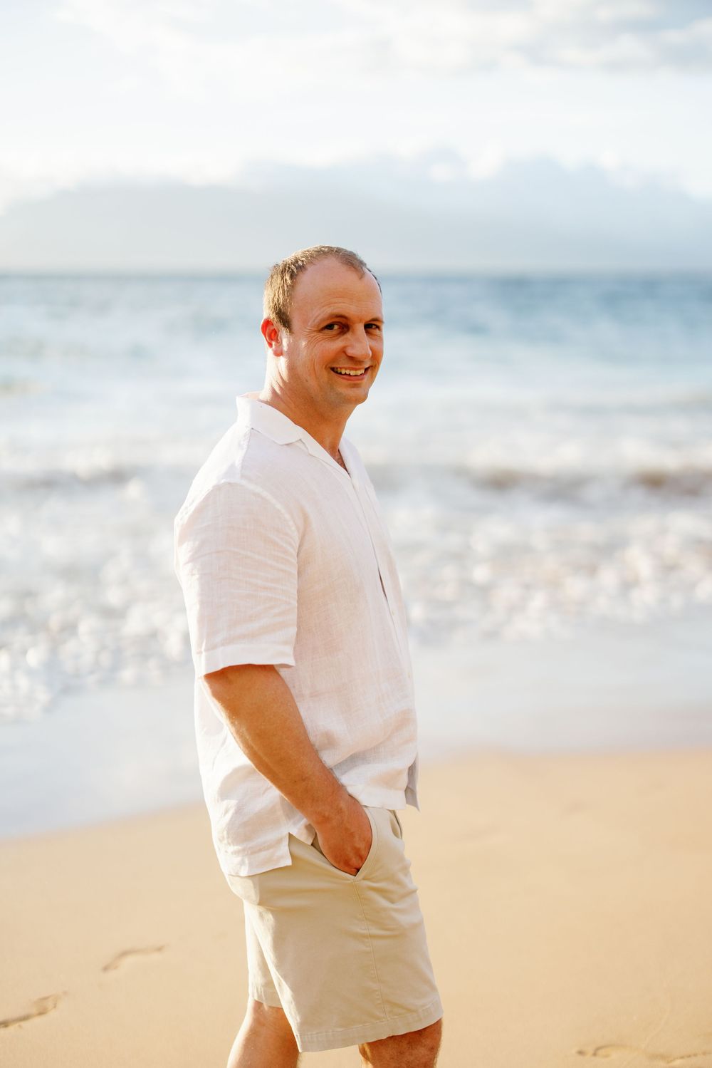 A smiling man in a white linen shirt and khaki shorts walks along a sunny sandy beach.