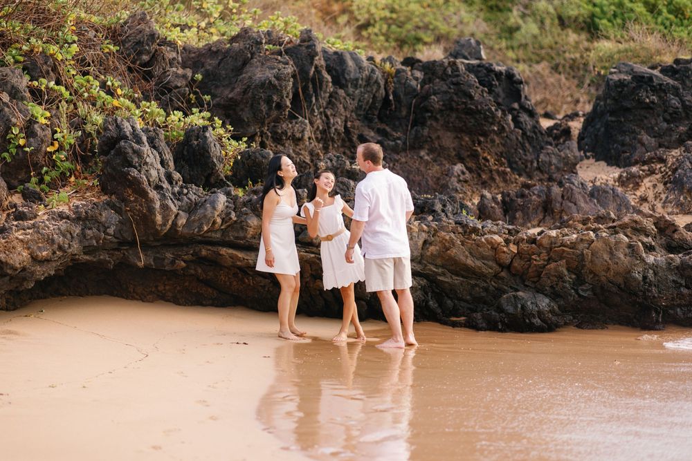 Family of three in white outfits standing barefoot on sandy beach near rocky cliffs.