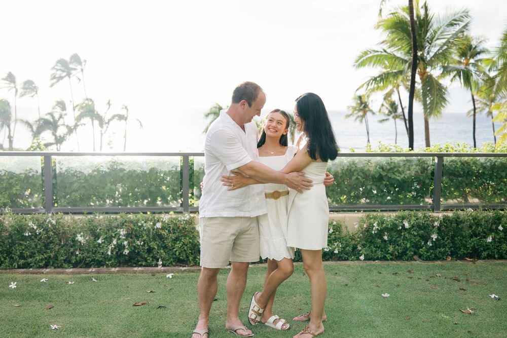 Family of three embracing and laughing together on a tropical lawn with palm trees and ocean views in Hawaii.