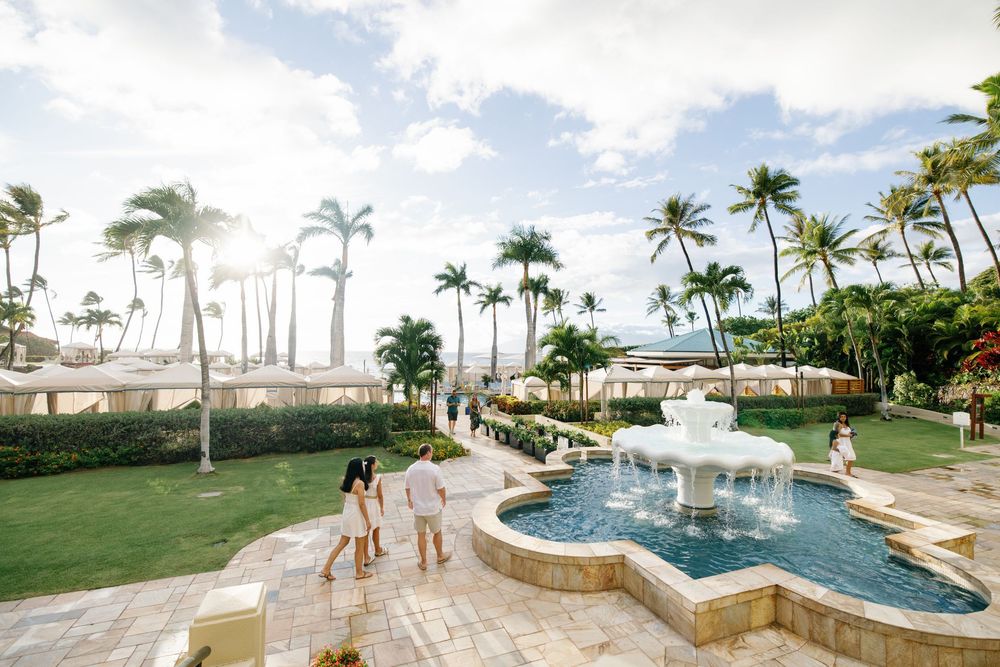 Couple strolling past a tropical resort fountain surrounded by lush palm trees and manicured gardens in Hawaii.