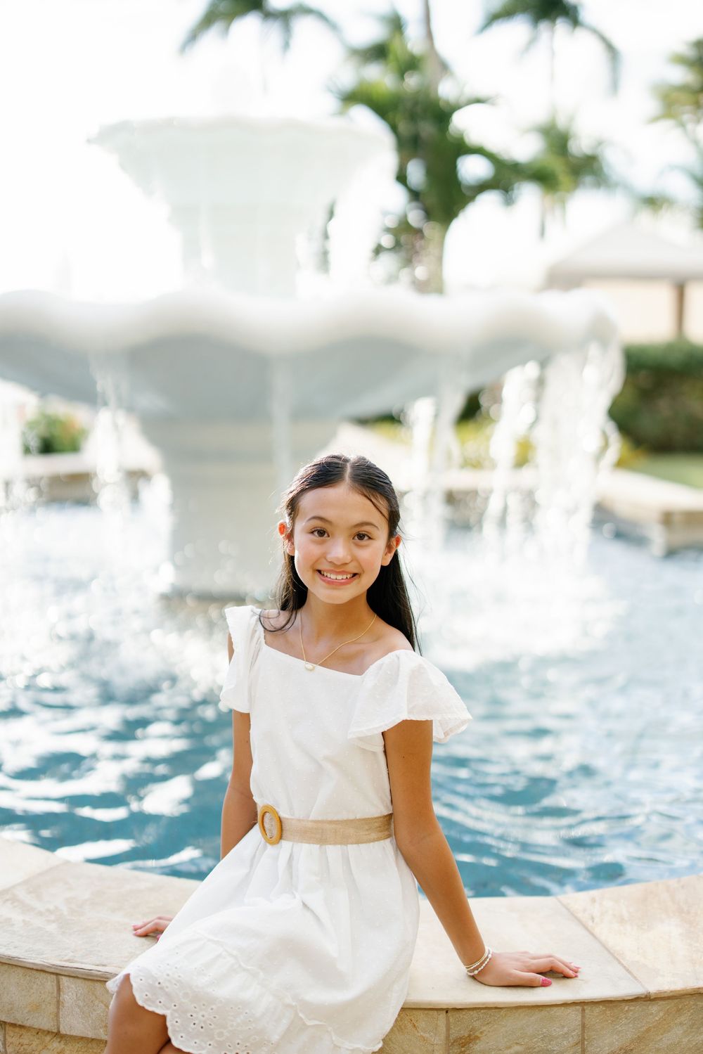 A smiling girl in a white dress with gold belt sits by an ornate fountain at a tropical resort.