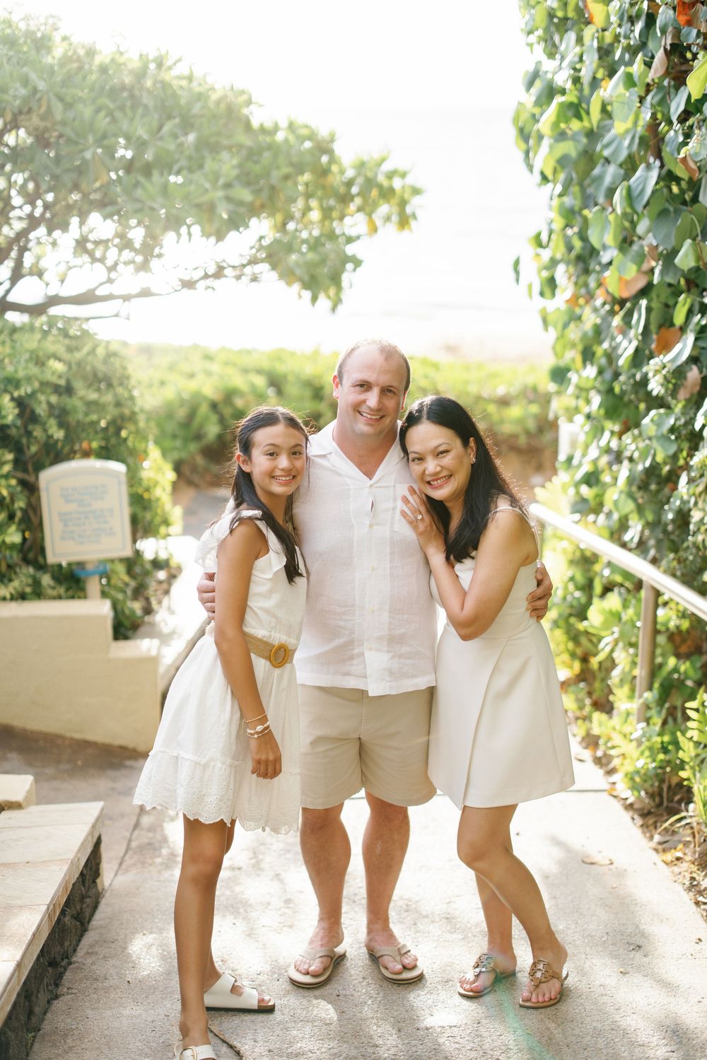 Family of three posing on a tropical pathway, wearing coordinated white outfits, smiling together on a sunny day.