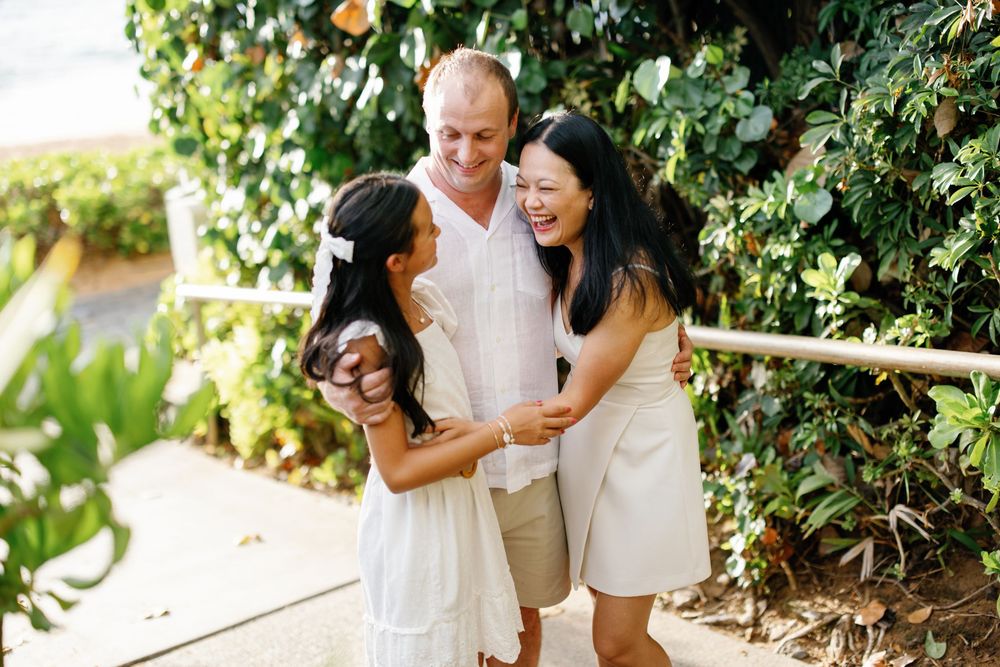 A joyful family of three laughing together outdoors surrounded by lush green tropical foliage.