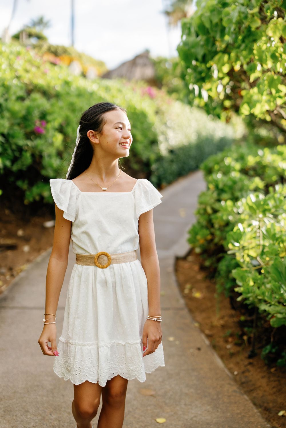 A smiling young girl in a white dress with a tan belt walks along a lush green garden path.