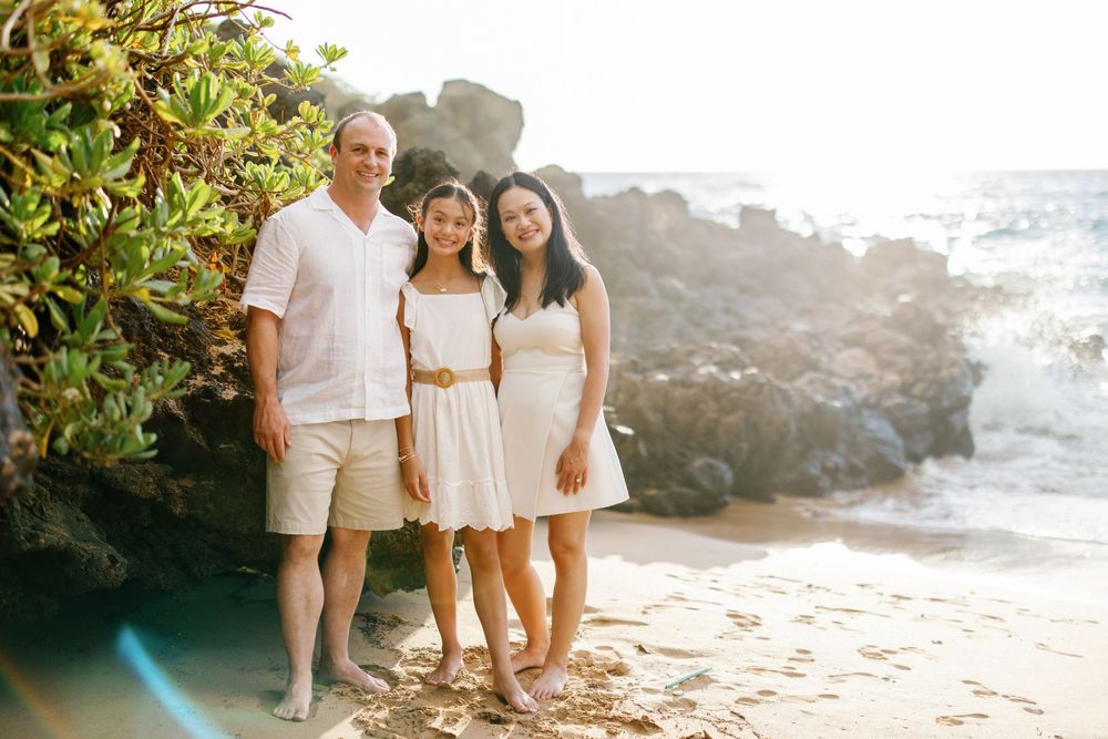 Family of three posing on a sandy beach in white outfits with rocky coastline and tropical greenery behind them.