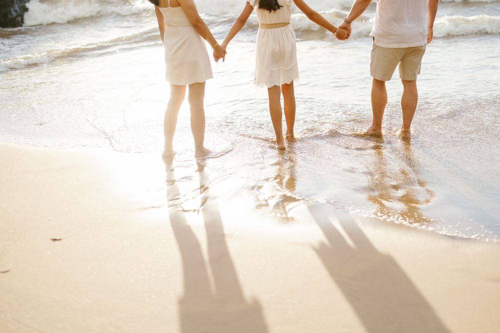 Family of three holding hands on a sunny beach, wearing white outfits, standing barefoot in ocean waves.