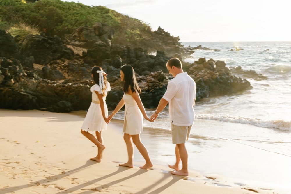Family of three in white outfits holding hands on a sunny sandy beach with rocky coastline in Hawaii.