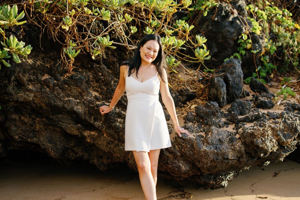 Young woman in white dress smiling on sandy beach with rocky cliffs and lush greenery backdrop.