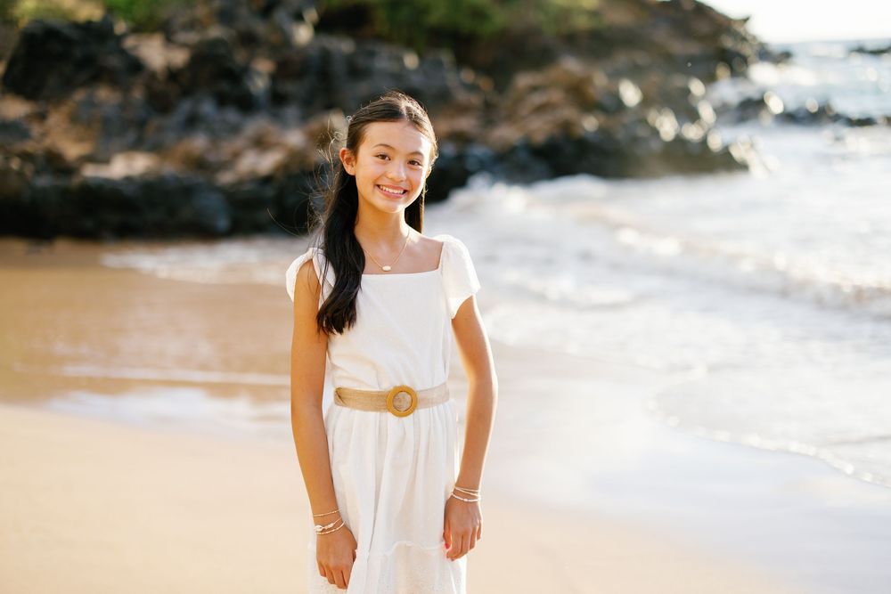 Young girl in white dress with gold belt smiling on sandy beach with ocean waves and rocks behind her.