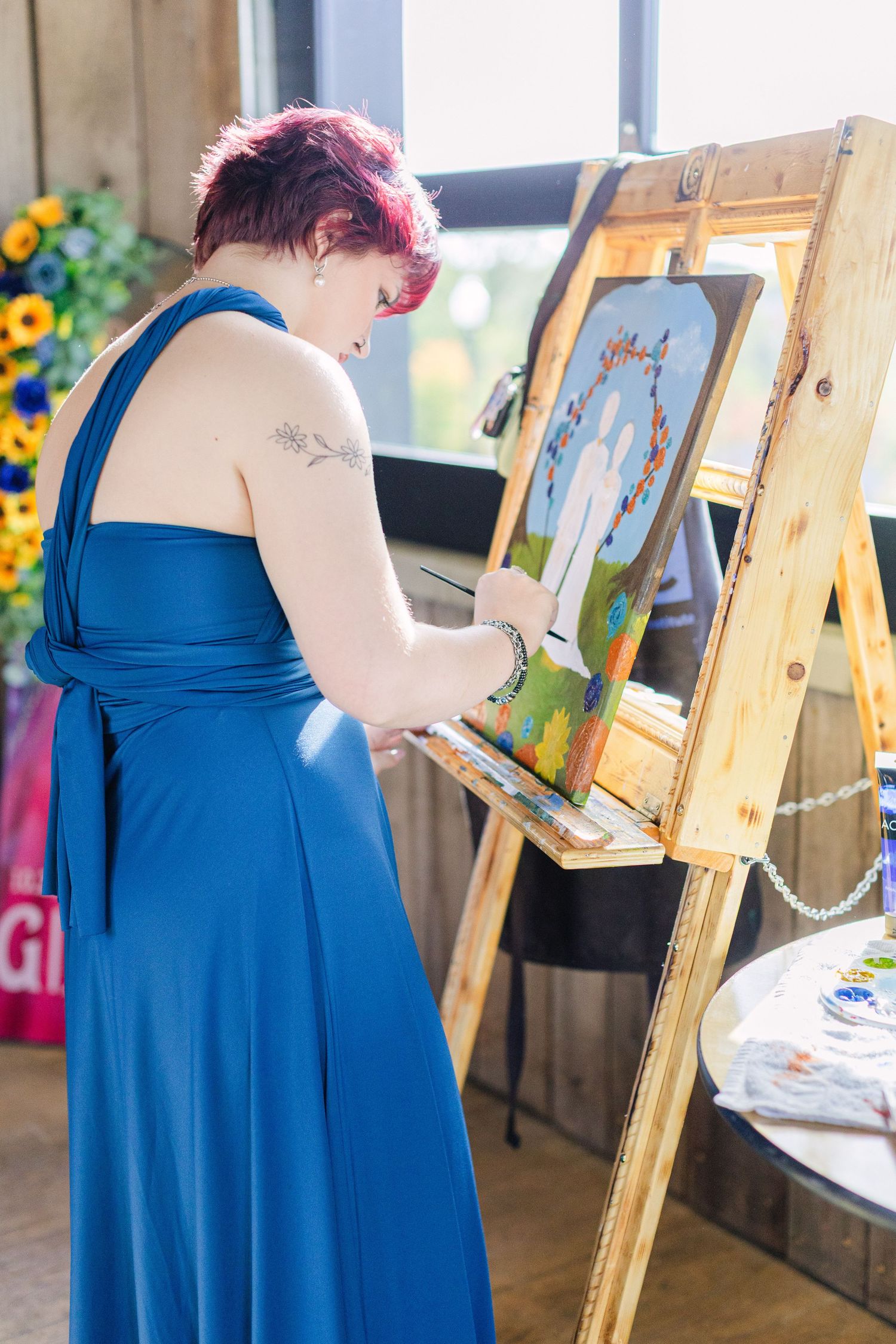 Woman in blue gown painting on easel at event, with red hair and tattoo, creating colorful artwork indoors.