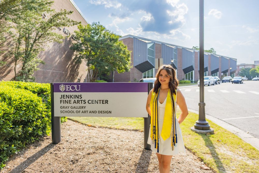 Graduate in cap and gown poses by ECU Jenkins Fine Arts Center sign on a sunny day.