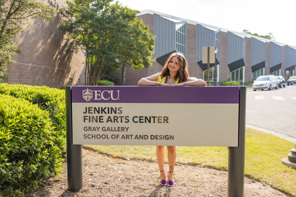 Woman posing beside the Jenkins Fine Arts Center sign at ECU Gray Gallery School of Art and Design.