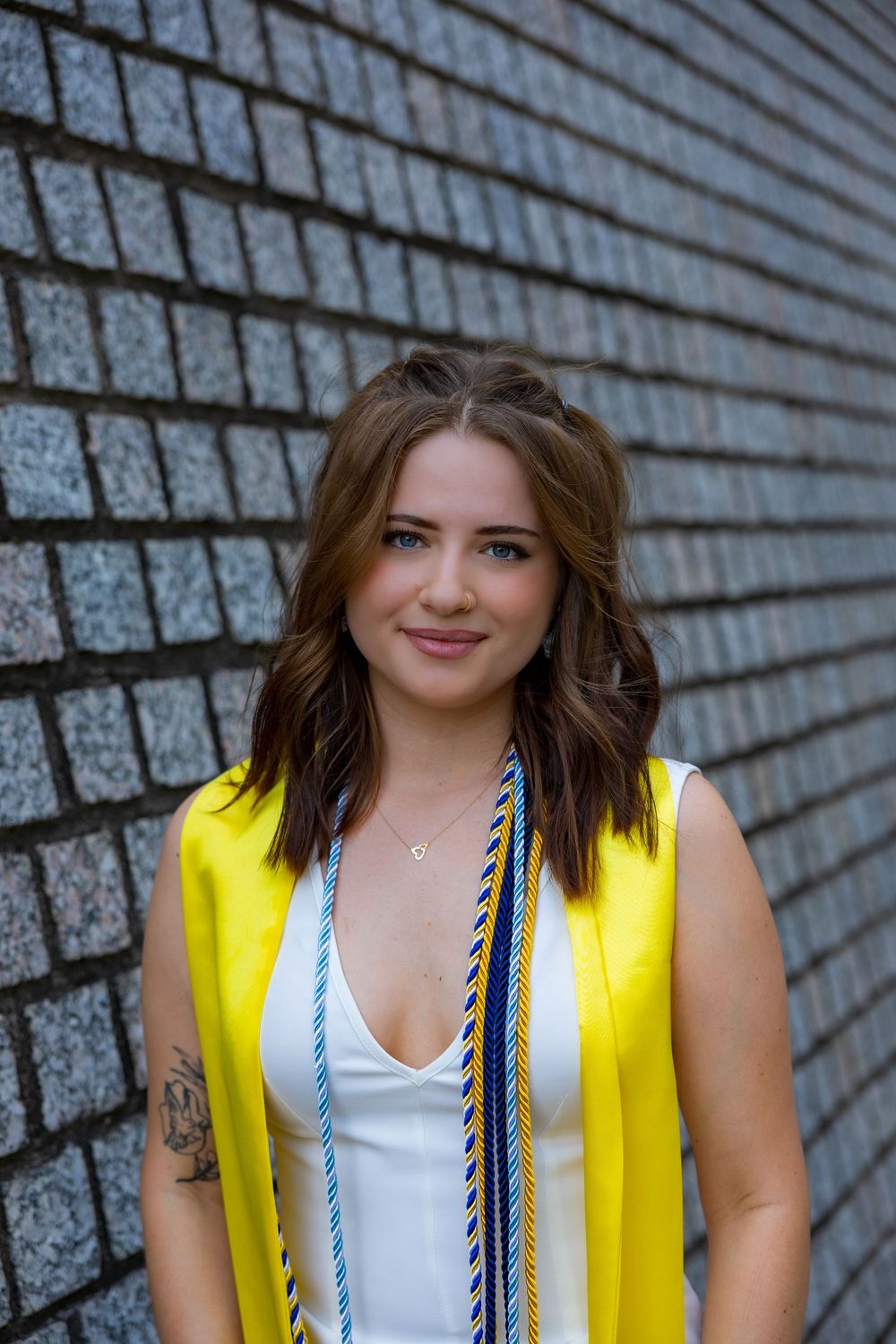 Young woman in yellow graduation stole and white top smiling against a brick wall backdrop.
