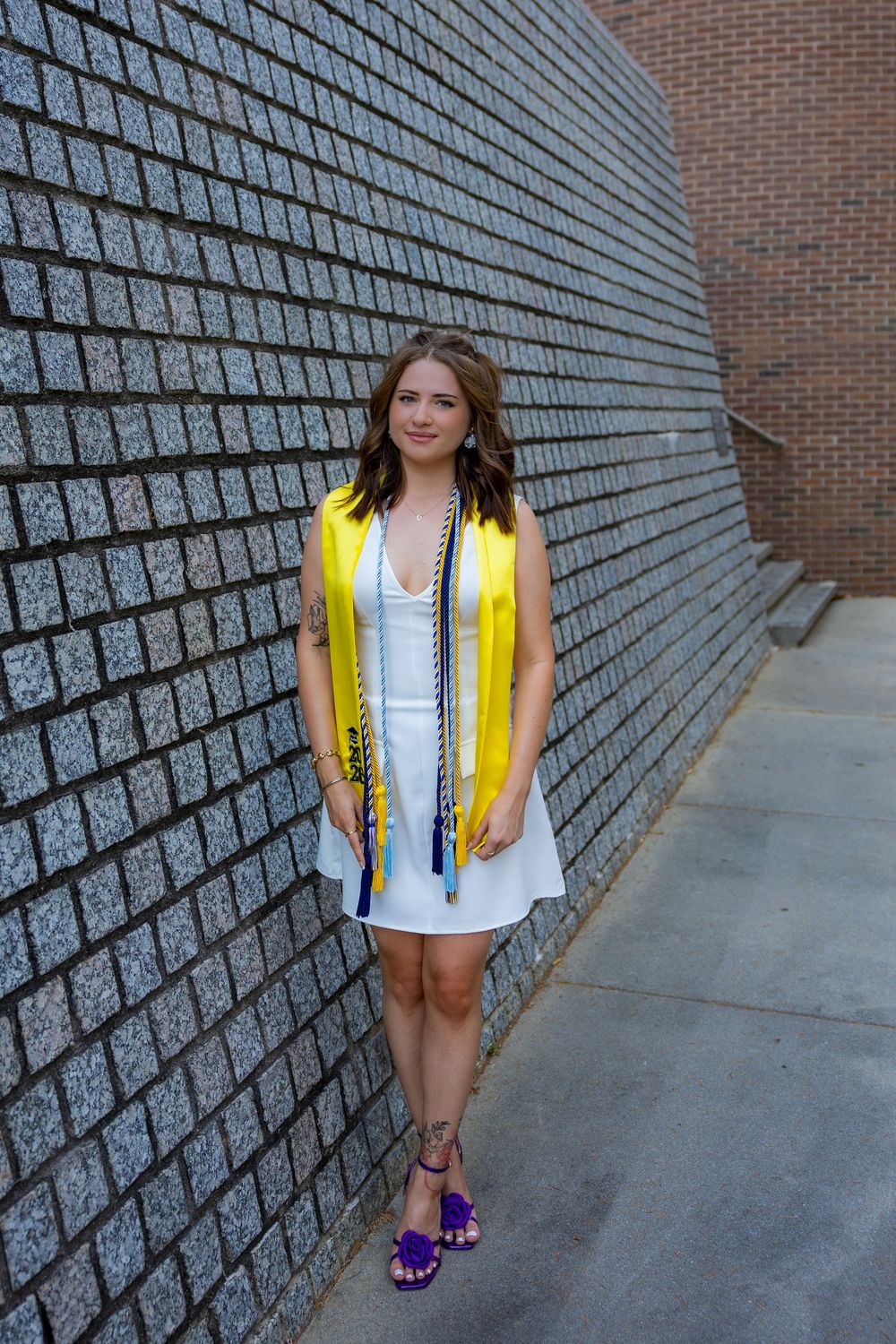 Young woman in white dress and yellow graduation stole poses against brick wall in purple sandals.