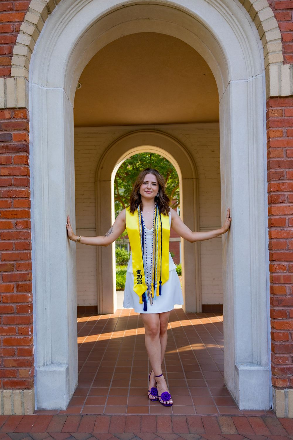 Young woman in white dress with yellow graduation stole poses under brick archway for graduation photos.