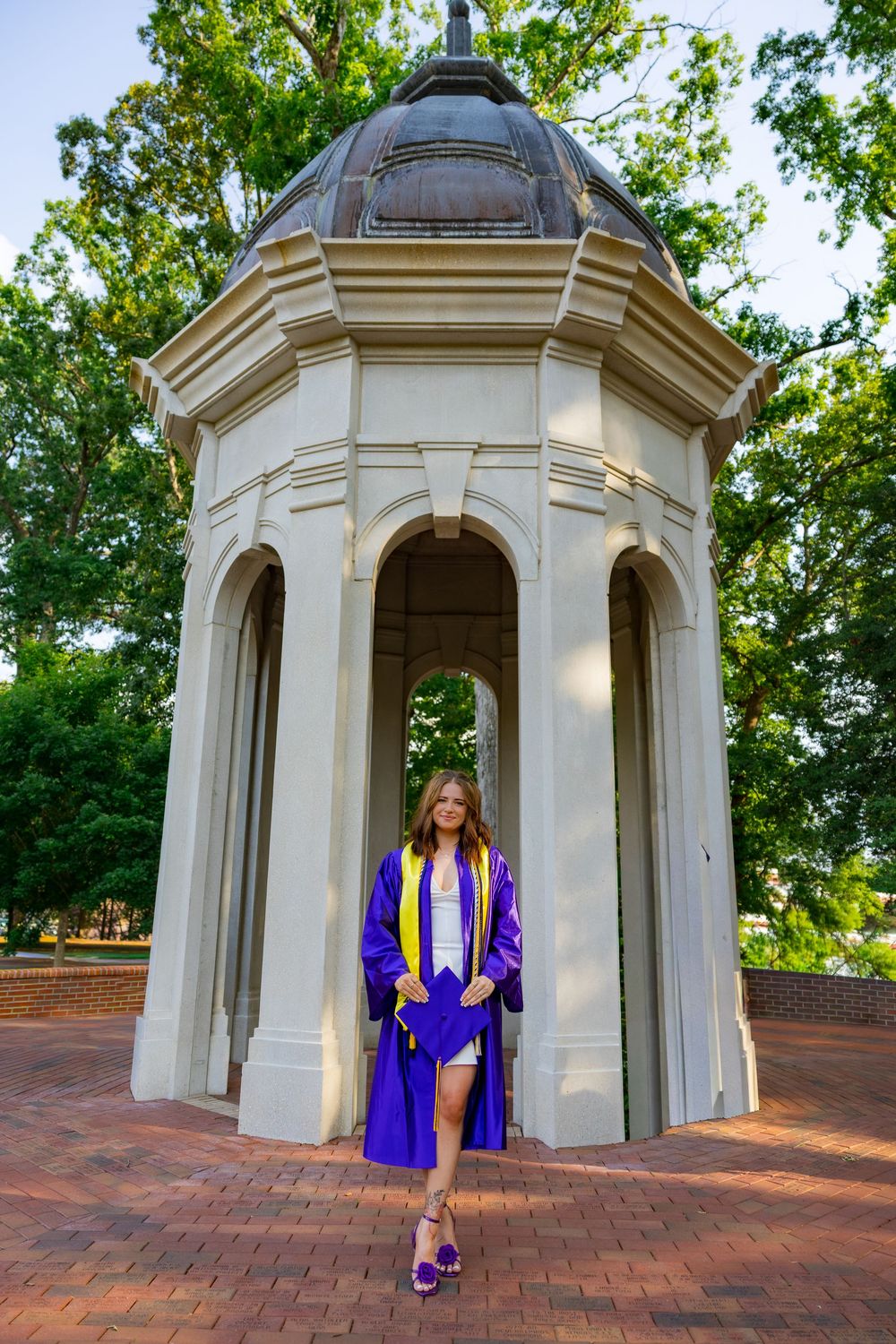 A smiling graduate in purple cap and gown poses in front of a white domed gazebo on a brick campus path.