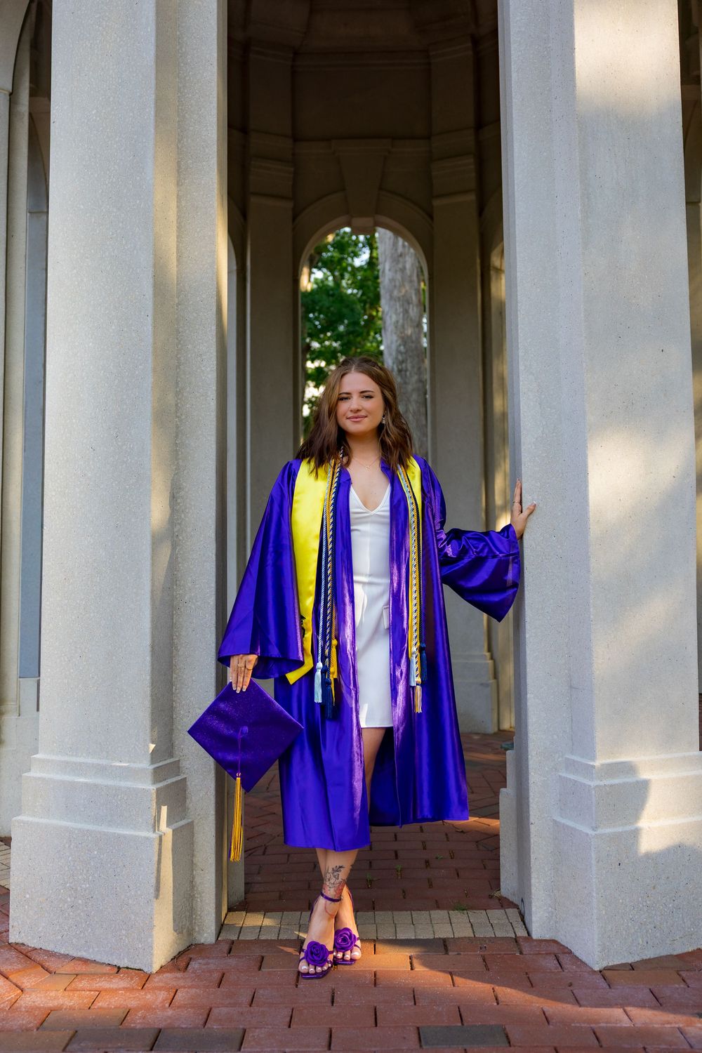 A smiling graduate poses in a purple cap and gown with yellow tassels between stone columns on brick pavement.