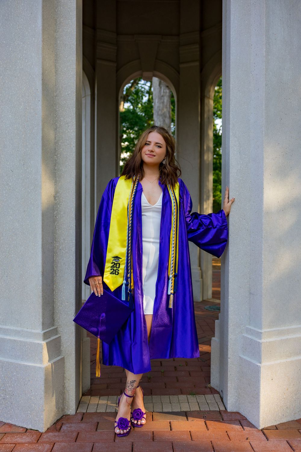 A young woman in a purple graduation gown and yellow sash poses between columns holding her cap.