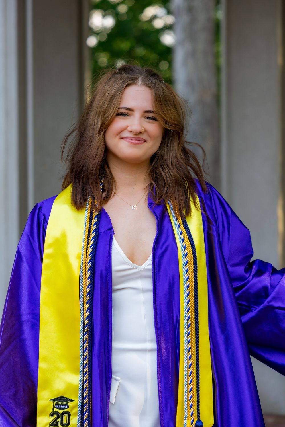 Young woman in purple graduation gown and yellow honor stole smiling outdoors.