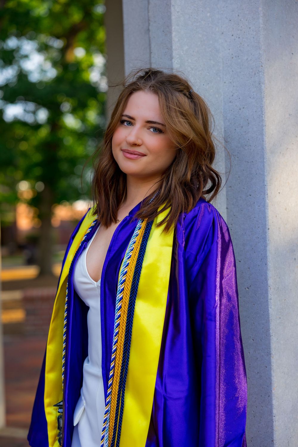 Young woman in blue and gold graduation gown smiles while leaning against a stone column outdoors.