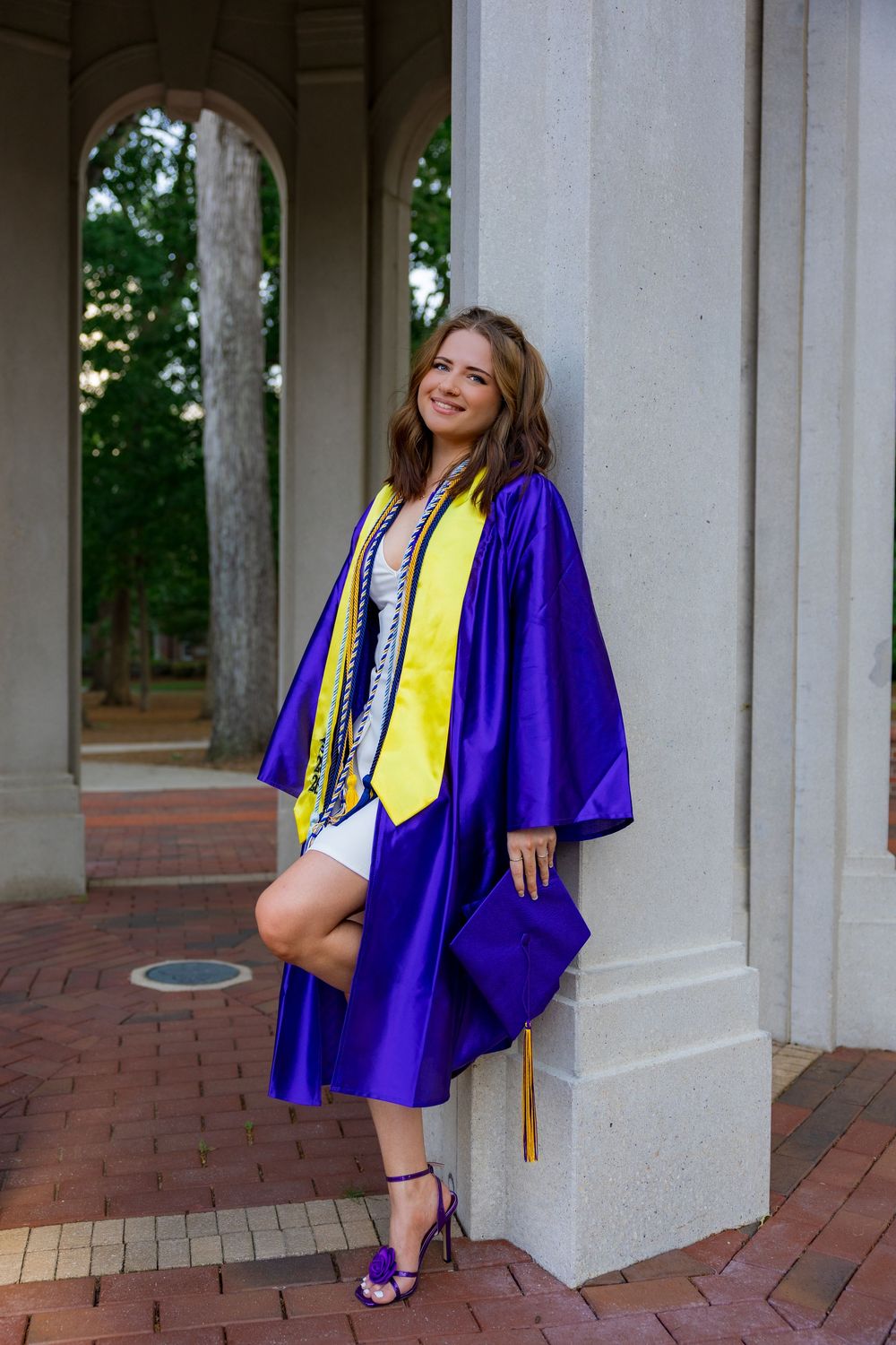 Young woman in purple graduation gown and yellow stole poses proudly against white column at outdoor ceremony venue.