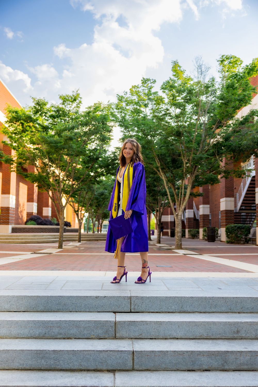 A young woman in purple graduation cap and gown stands proudly on steps outside a brick university building.