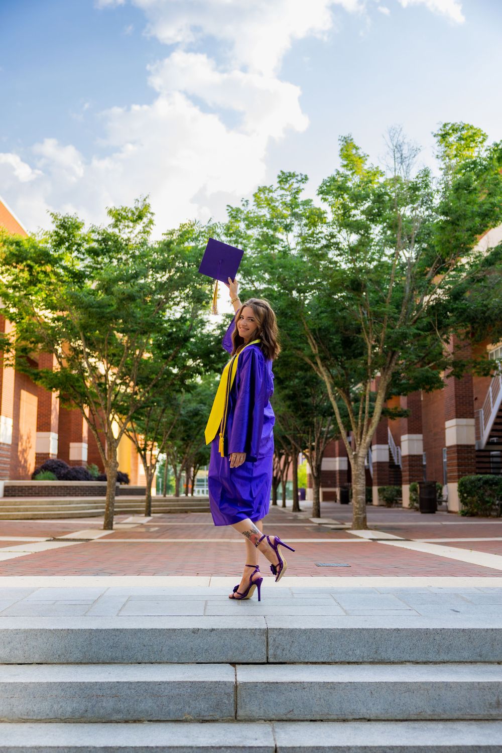 A joyful graduate in purple cap and gown tosses her mortarboard on a sunny campus walkway lined with trees.