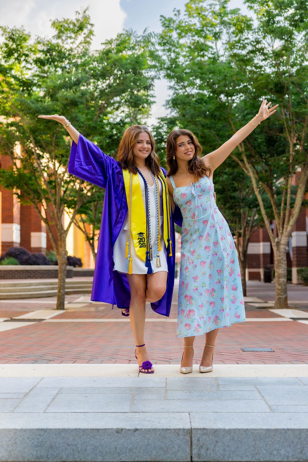 Two young women celebrating graduation, one in purple cap and gown, posing joyfully outdoors on a brick plaza.